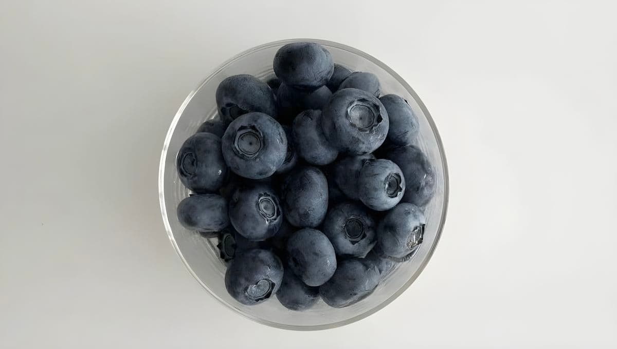 A clear glass bowl of fresh blueberries seen from above on a white background